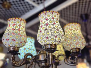 A stunning chandelier, its metallic frame supporting a collection of lampshades. Each shade is a masterpiece of mosaic art, showcasing delicate white flowers with colorful centers. A touch of luxury and elegance. It was spotted in an electrical shop in Kozhikode, Kerala.