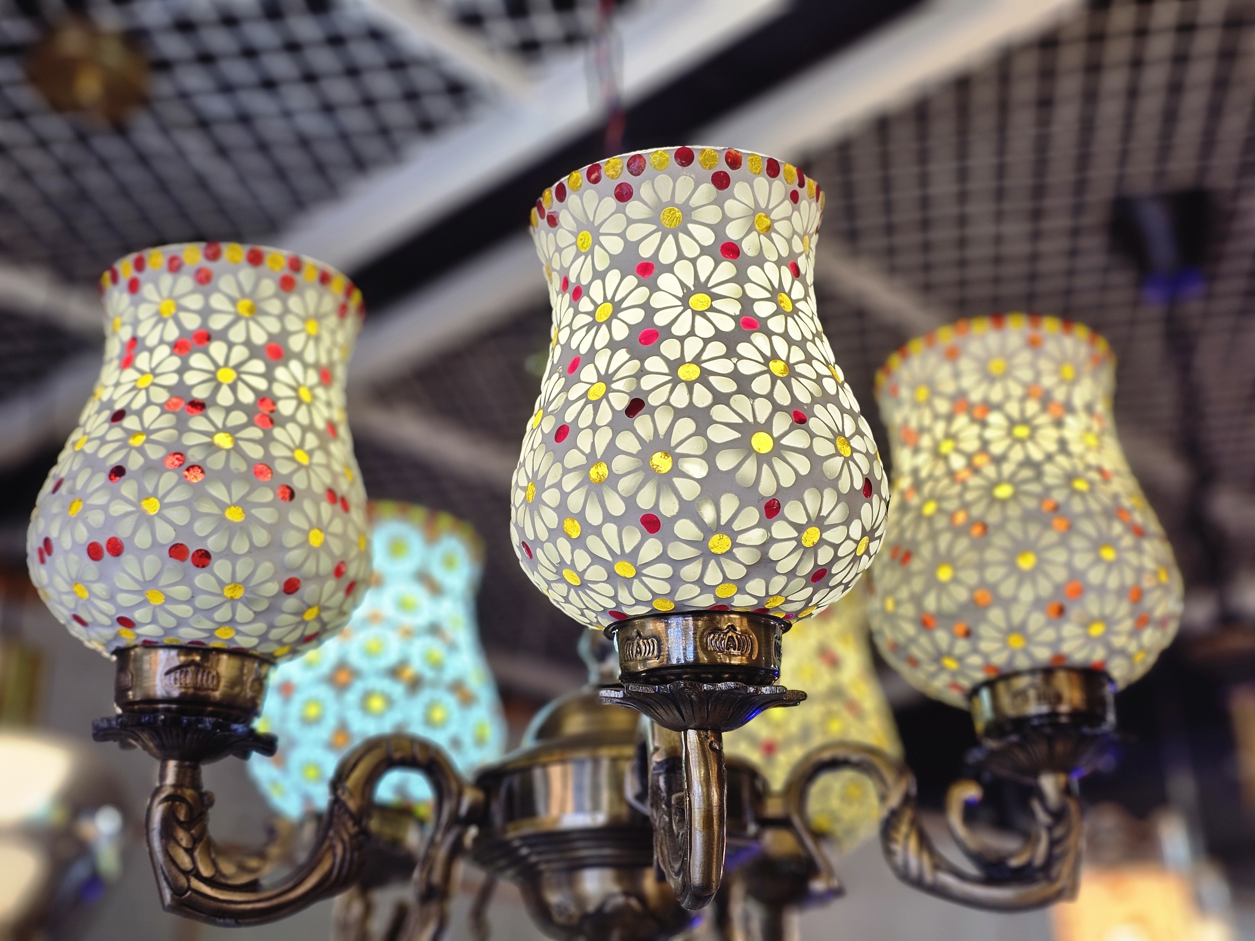A stunning chandelier, its metallic frame supporting a collection of lampshades. Each shade is a masterpiece of mosaic art, showcasing delicate white flowers with colorful centers. A touch of luxury and elegance. It was spotted in an electrical shop in Kozhikode, Kerala.