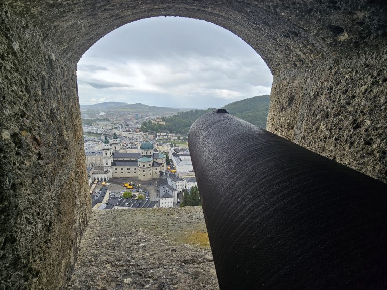 A view from Fortress Hohensalzburg, Salzburg, with a cannon in the foreground and the city of Salzburg in the background. The view is framed by a stone archway. The sky is cloudy, and mountains are visible in the distance.