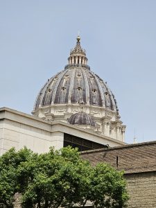 The majestic dome of St. Peter's Basilica (Vatican) towers above the surrounding buildings and trees, its intricate stonework and cross gleaming against a clear sky. Partially hidden below, a mix of modern and historic architecture frames the scene, blending past and present. 