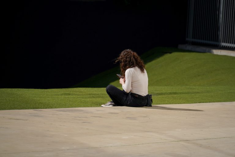 A person with curly hair wearing a white long-sleeve shirt and black pants is sitting on a grassy area, using a smartphone. The ground is partly concrete, and there is a dark wall and railing in the background.