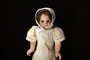 A vintage doll wearing a white bonnet and a cream-colored dress, set against a black background.