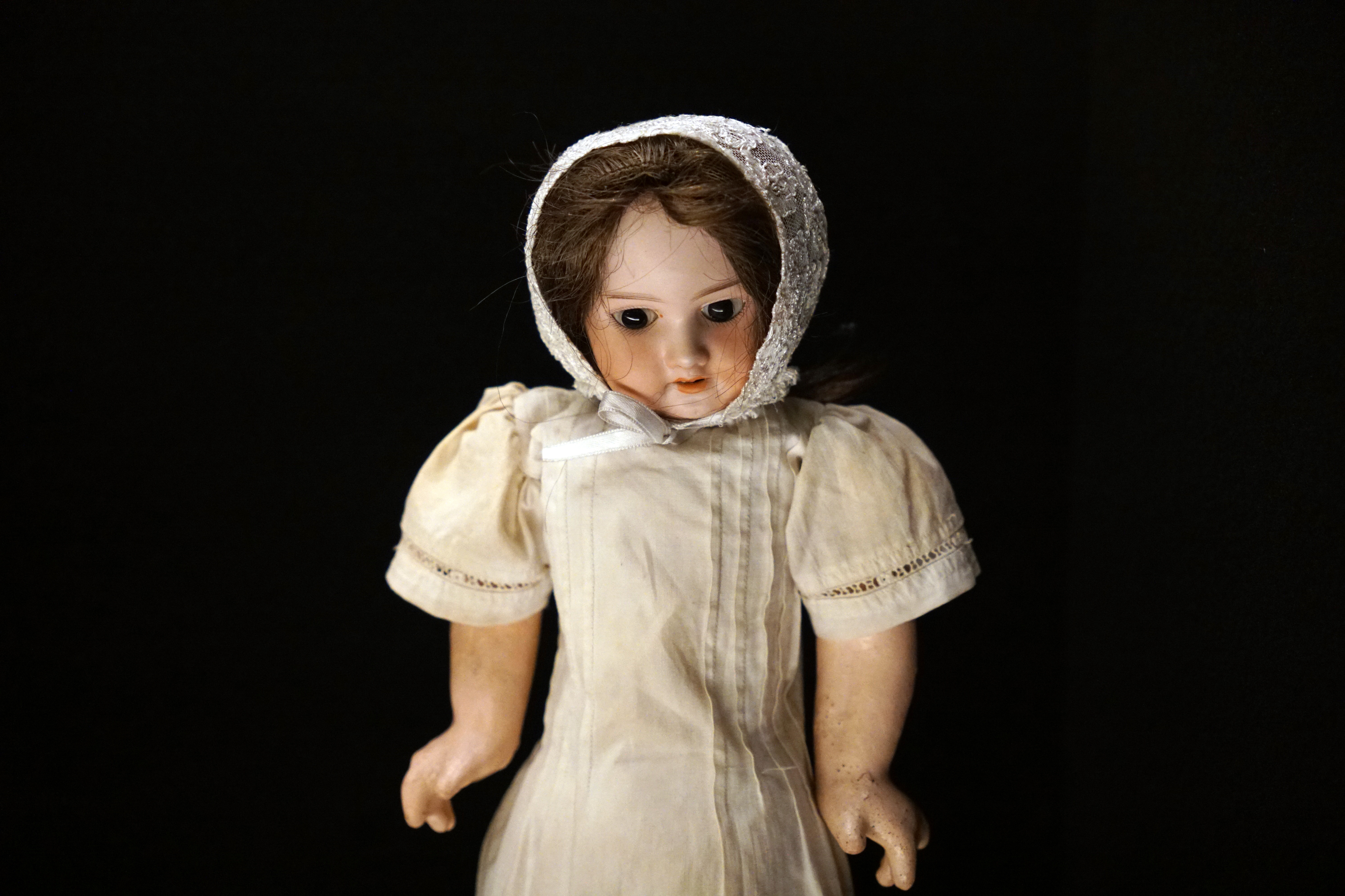 A vintage doll wearing a white bonnet and a cream-colored dress, set against a black background.