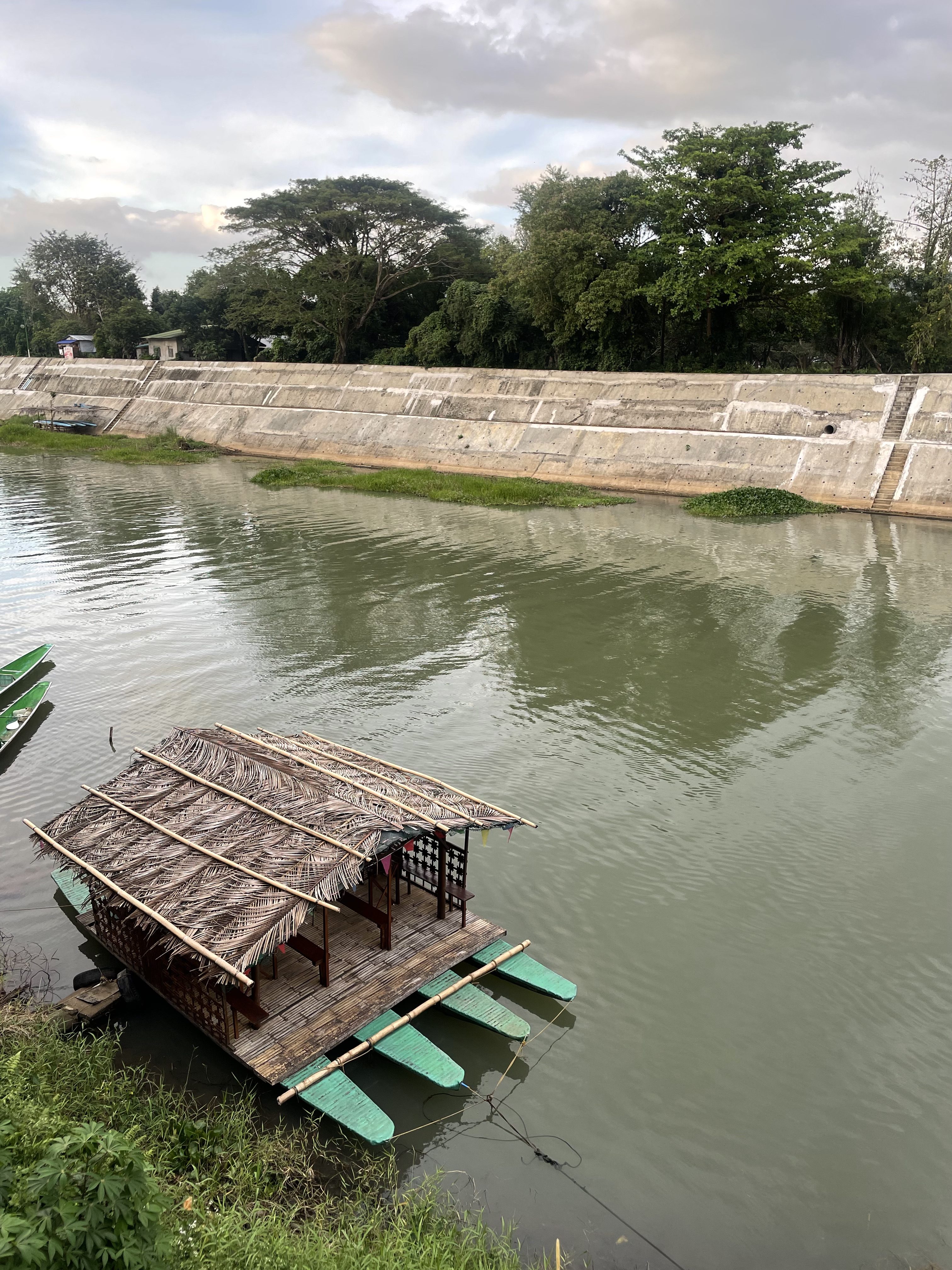 A bamboo raft with a thatched roof is floating on a calm river. The riverbank has a concrete wall and is lined with lush green trees.