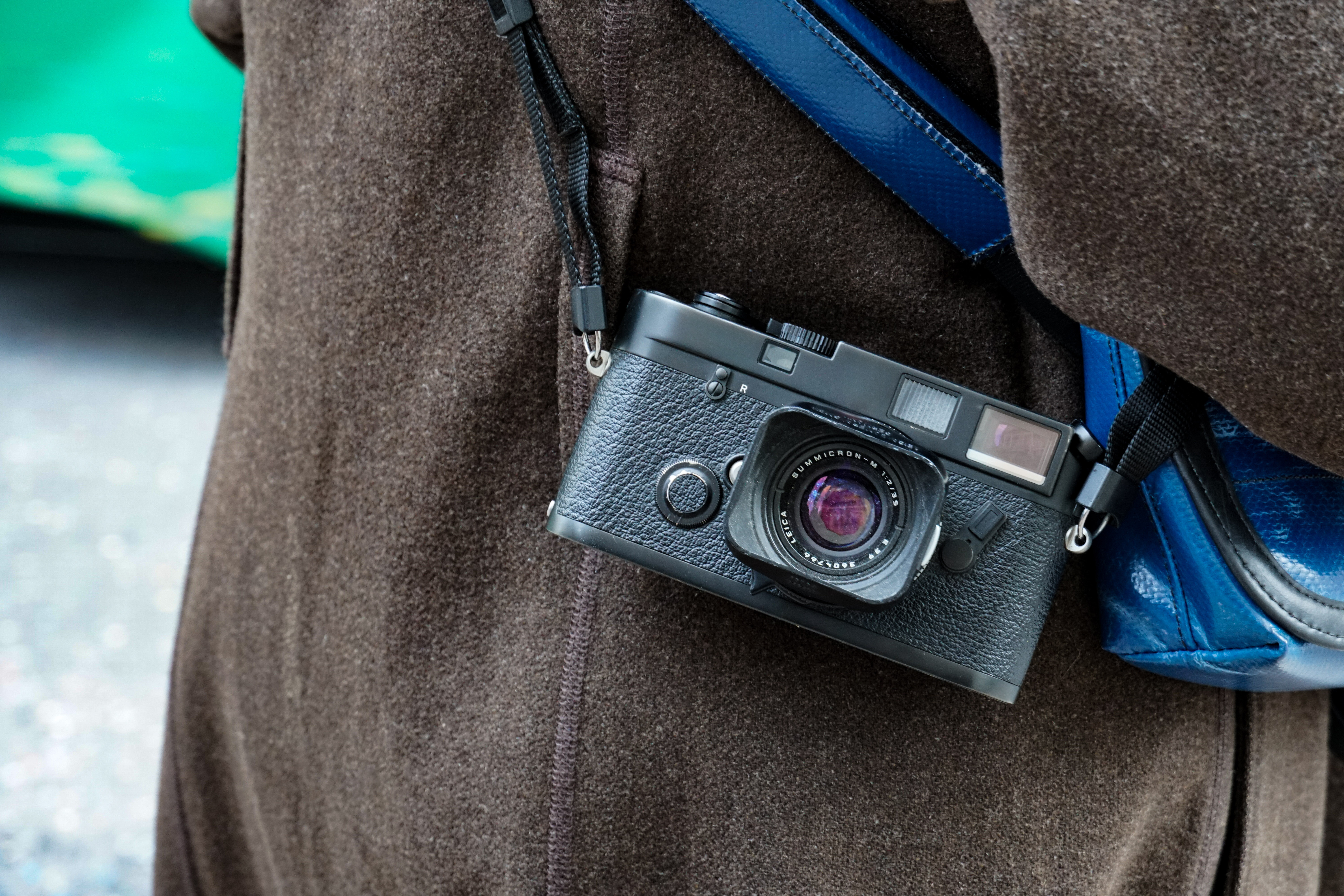 A close-up of a black rangefinder camera hanging from a strap against a person wearing a brown jacket