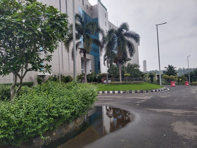 A modern building with a glass facade surrounded by lush greenery and palm trees. There is a clear sky in the background, and a wet pathway with a puddle in the foreground reflecting part of the building and trees.