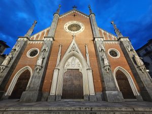 The facade of Santa Giulia church in Turin, Italy, at night. The church features a red brick facade with grey stone accents, statues, and decorative elements. The central wooden doors are flanked by arched entrances, and a rose window is visible above, all set against a cloudy evening sky.

