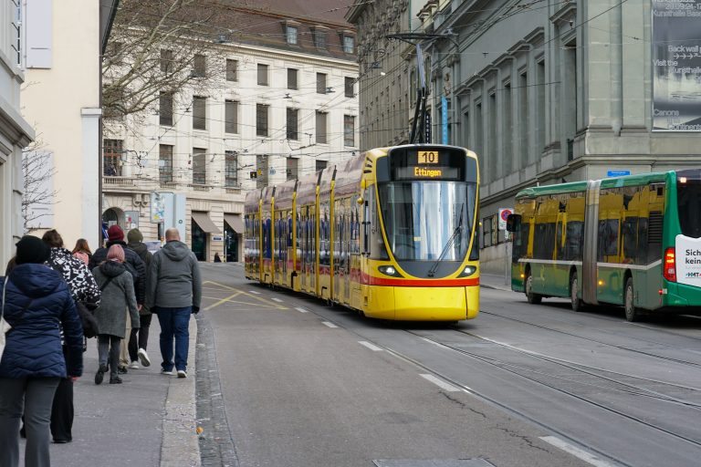 A yellow tram numbered 10 is traveling on a city street with buildings in the background. A green bus is visible alongside the tram. A group of people are walking on the sidewalk.