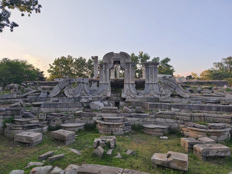 Ruins of an ancient structure with ornate stone carvings and pillars, surrounded by scattered pieces of masonry on grass, set against a backdrop of trees and a clear sky.