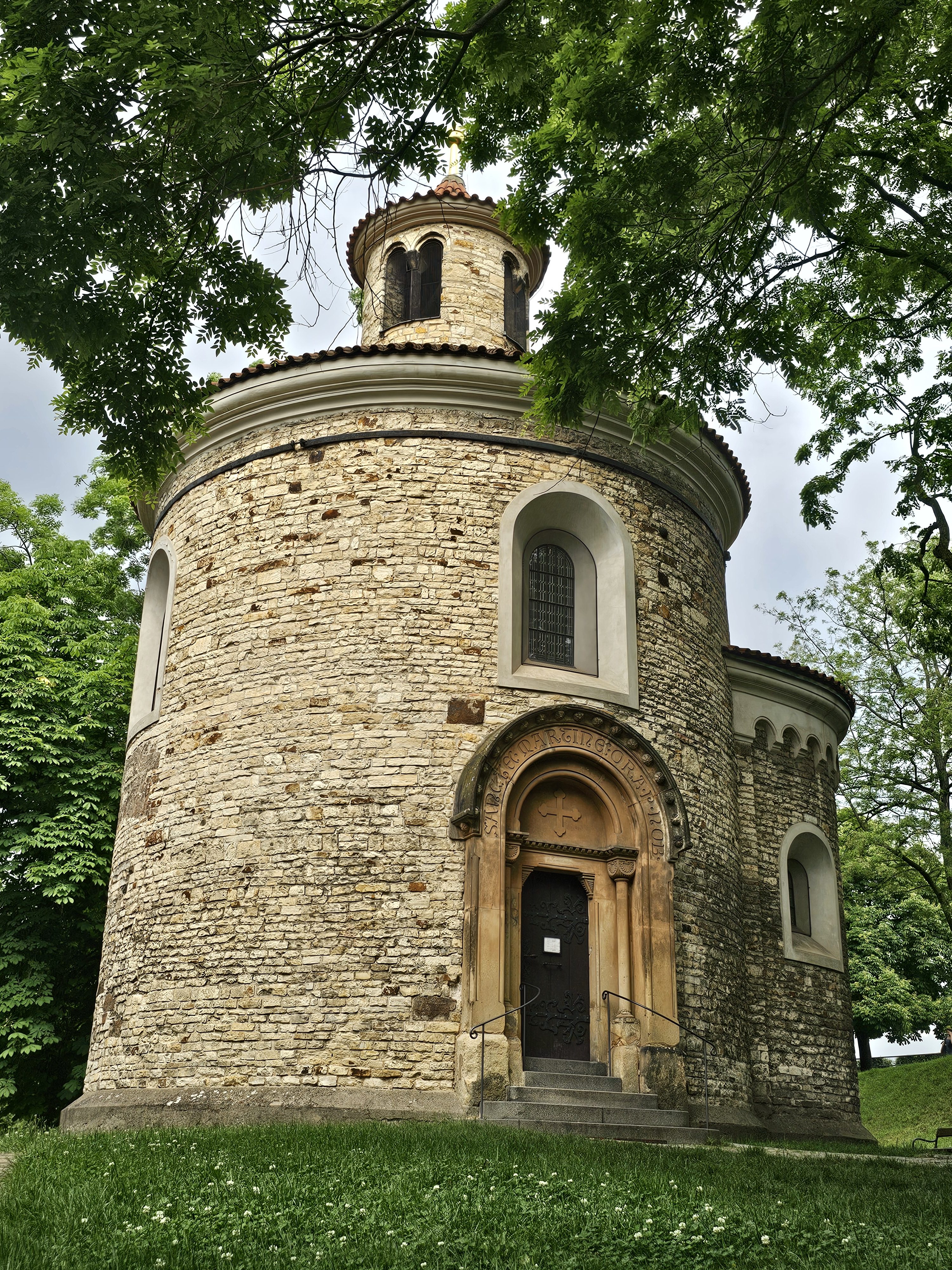 The Rotunda of St. Martin, a Romanesque church located in Vyšehrad, Prague. The rotunda is a cylindrical stone building with a conical roof and small windows. It is surrounded by trees and grass.