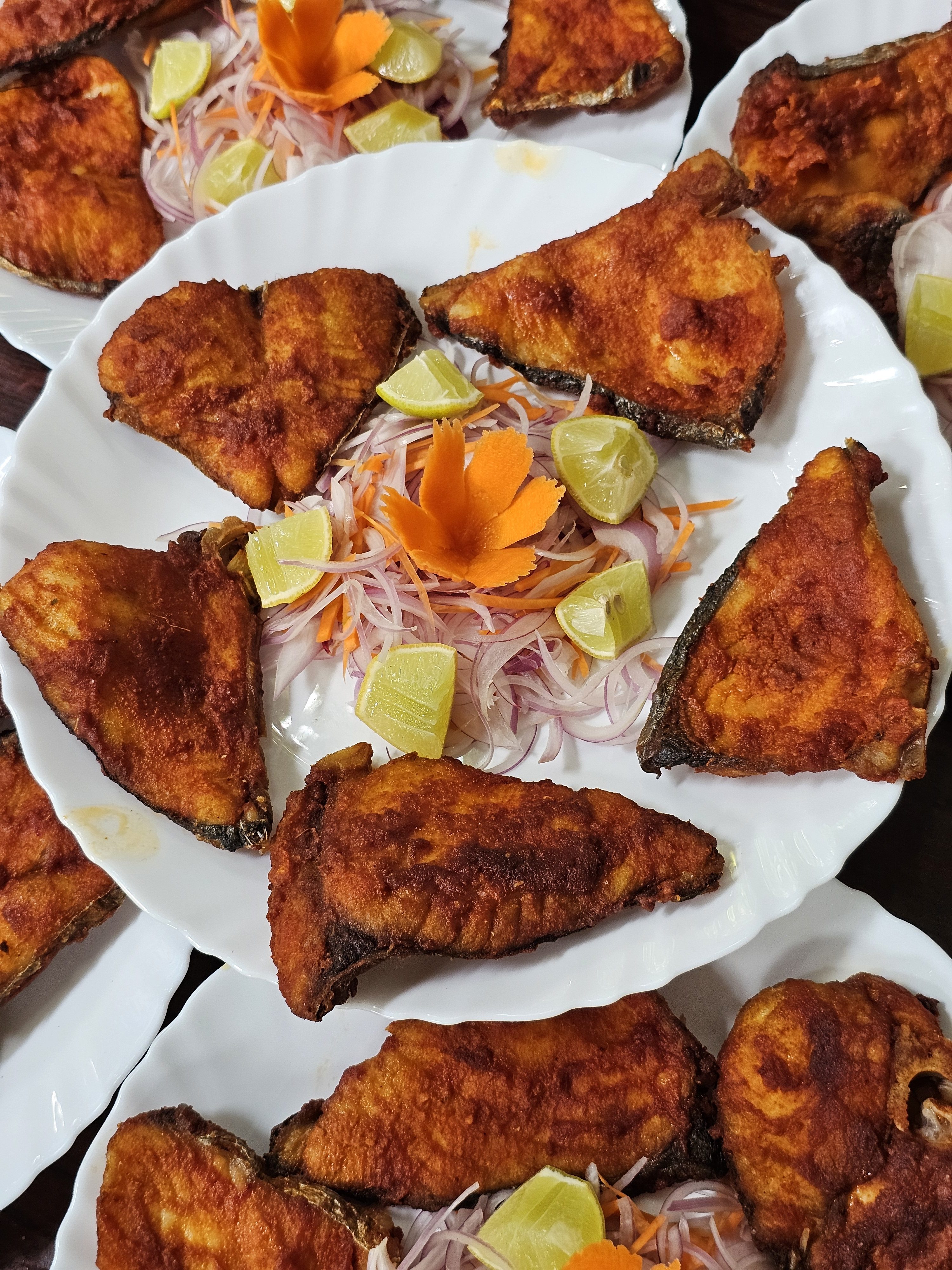 Multiple plates of fried fish (Narrow-barred Spanish mackerel), garnished with sliced onions, carrots, and lemon wedges. Each plate contains several pieces of crispy, golden-brown fish, they are ready to be served. Spotted in Kozhikode, Kerala.