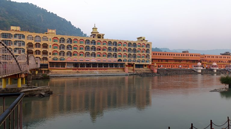 A large,haridwar building with colorful balconies stands by a calm river. A pedestrian bridge extends over the water, leading to the building. The background features a forested hillside under a clear sky.