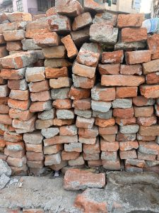 A stack of old, weathered red bricks with some gray mortar remnants, piled haphazardly. The background includes parts of buildings and a plastic bottle is visible on top right.
