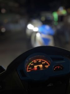 A close-up of a scooter dashboard showing an illuminated speedometer and fuel gauge. The background is blurred.