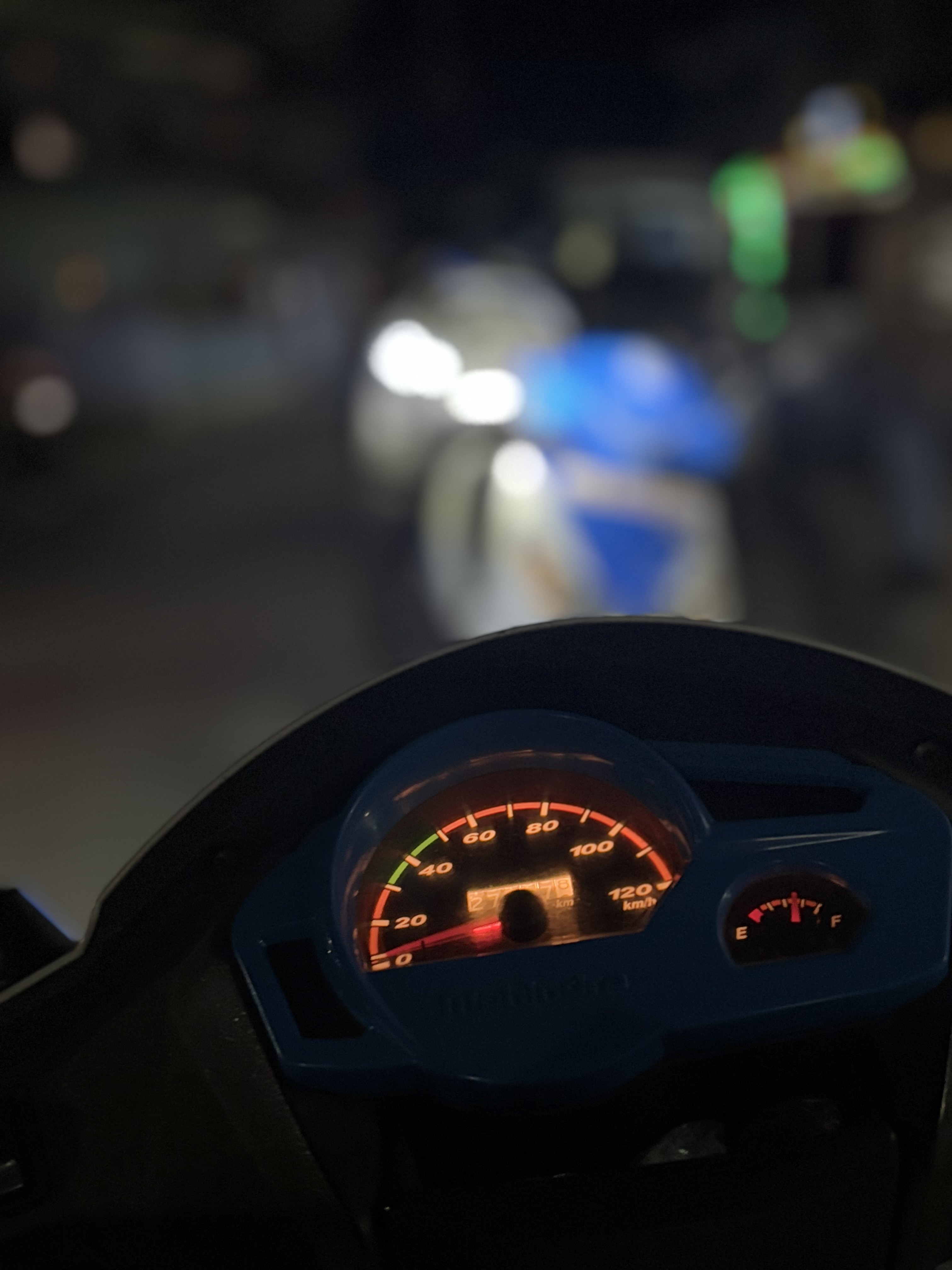 A close-up of a scooter dashboard showing an illuminated speedometer and fuel gauge. The background is blurred.