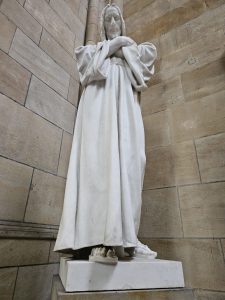 A full-body marble statue of Jesus Christ, standing on a white pedestal. The statue is positioned against a stone wall with visible seams and textures. Captured from St. Vitus Cathedral, Prague Castle. 