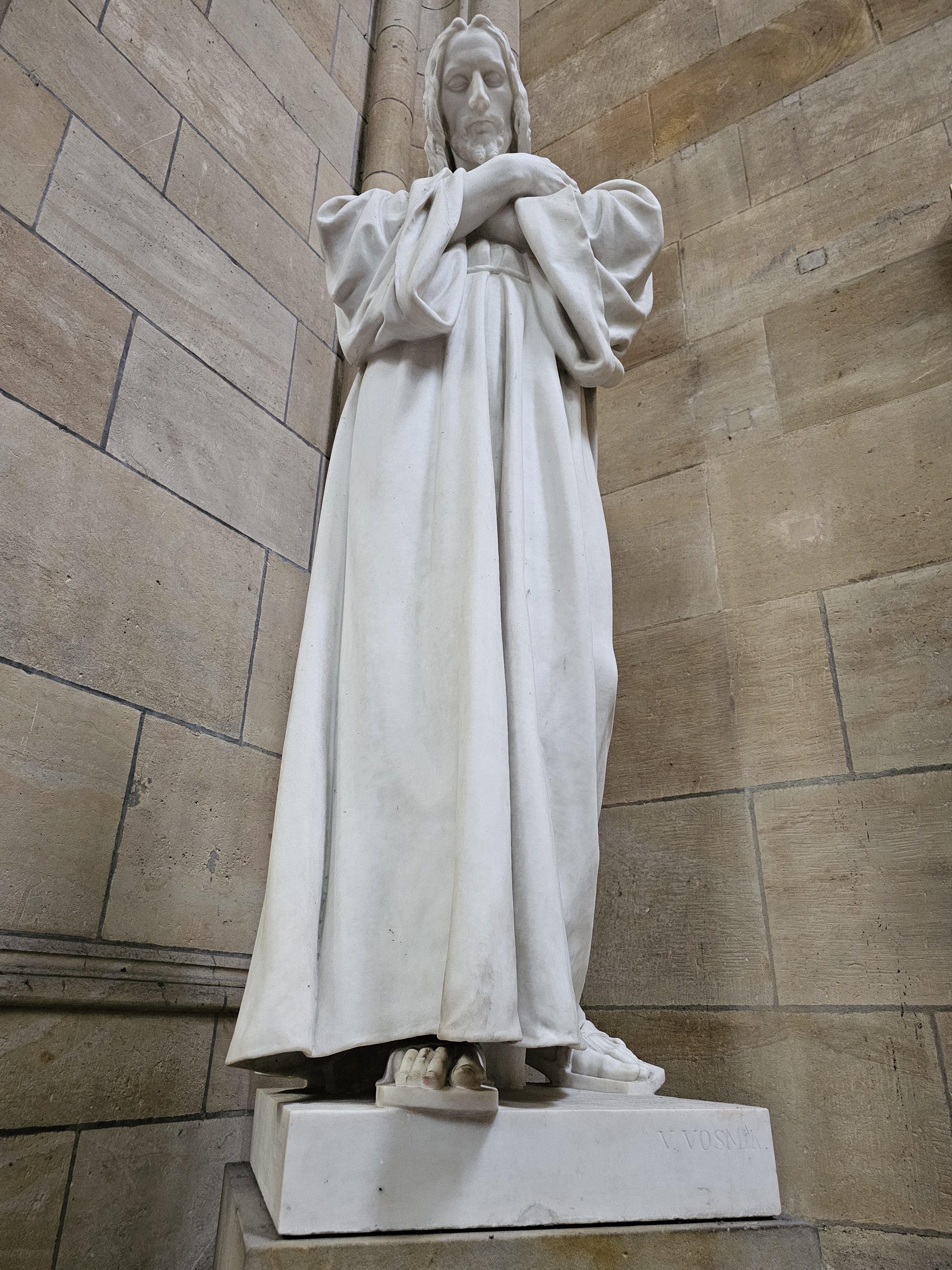 A full-body marble statue of Jesus Christ, standing on a white pedestal. The statue is positioned against a stone wall with visible seams and textures. Captured from St. Vitus Cathedral, Prague Castle.