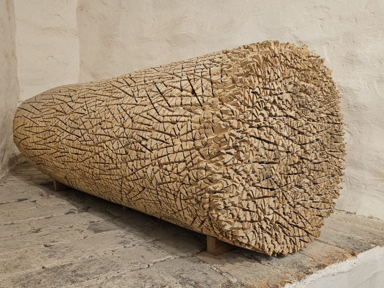 A light brown, cylindrical wood sculpture with a rough, textured surface. The sculpture is positioned horizontally, resting on two small wooden supports, and is set against a backdrop of a textured, off-white wall and a stone-like floor, from Hohensalzburg Fortress, Salzburg.