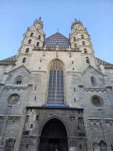 The facade of St. Stephen's Cathedral in Vienna, Austria, featuring its two towers and patterned roof. The Gothic architecture is captured from a low angle, emphasizing its height and detail against a clear sky. The cathedral's stone exterior and arched entrance are prominently displayed.
