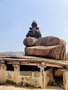 Ancient stone structure (Stone temple) with a flat roof supported by stone columns, topped by a triangular rock formation, set against a clear blue sky.Photo taken at Hampi, India.
