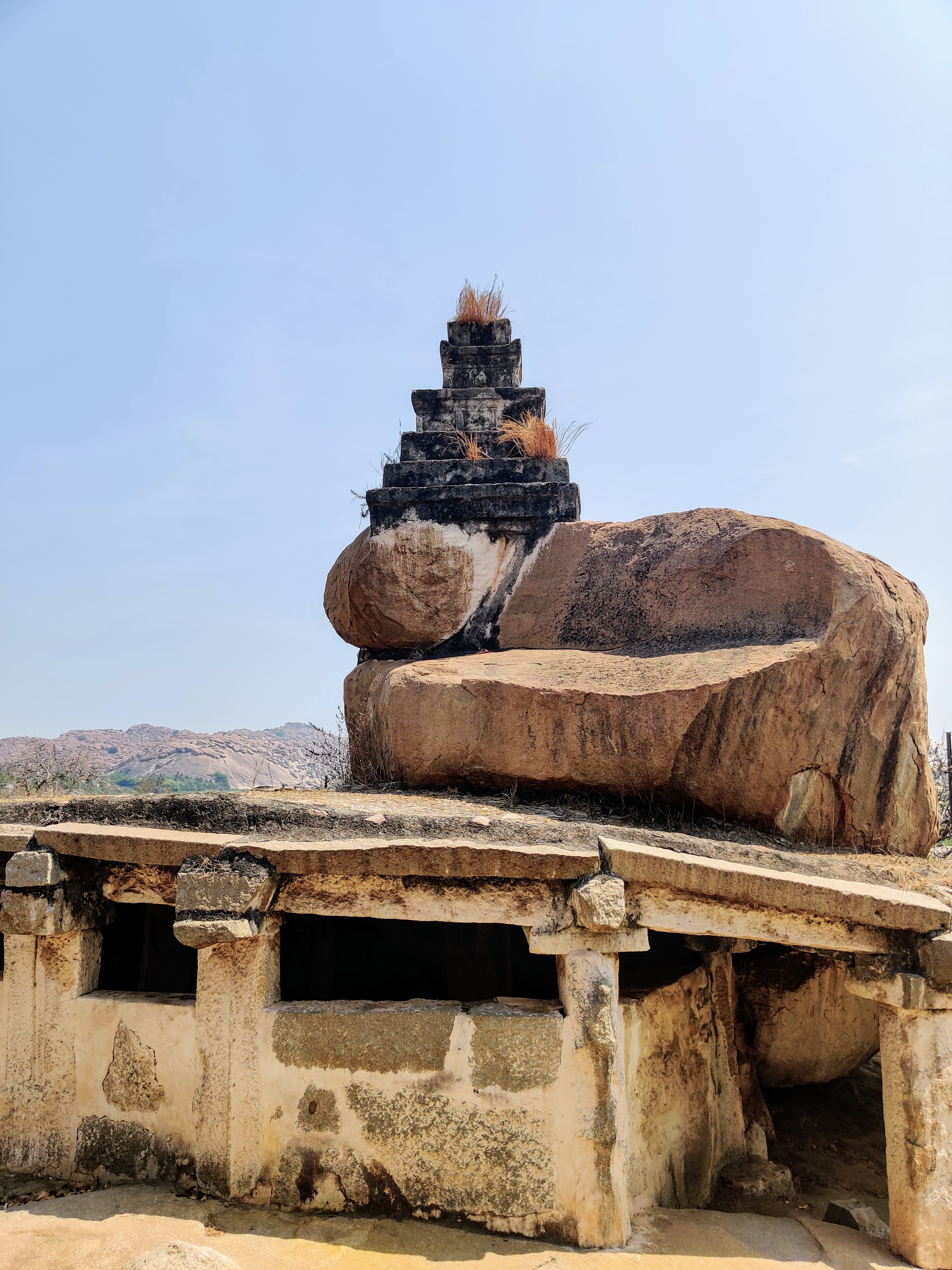 Ancient stone structure (Stone temple) with a flat roof supported by stone columns, topped by a triangular rock formation, set against a clear blue sky.Photo taken at Hampi, India.