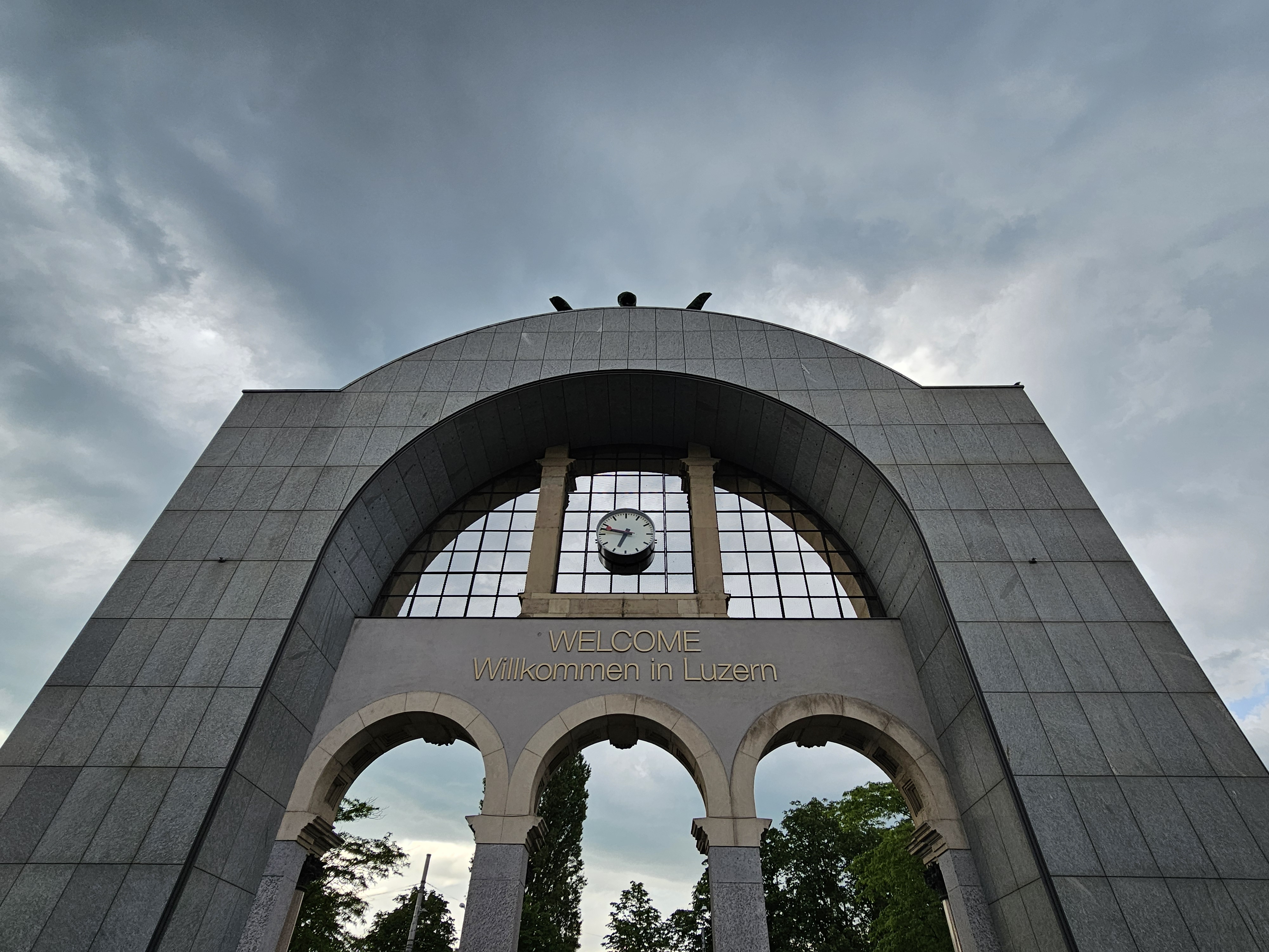 The entrance to Lucerne train station in Switzerland, features a large arched stone facade with the words "WELCOME Willkommen in Luzern" inscribed above three arched entrances. A clock is visible through the glass above the inscription, and the structure is set against a cloudy dark sky.