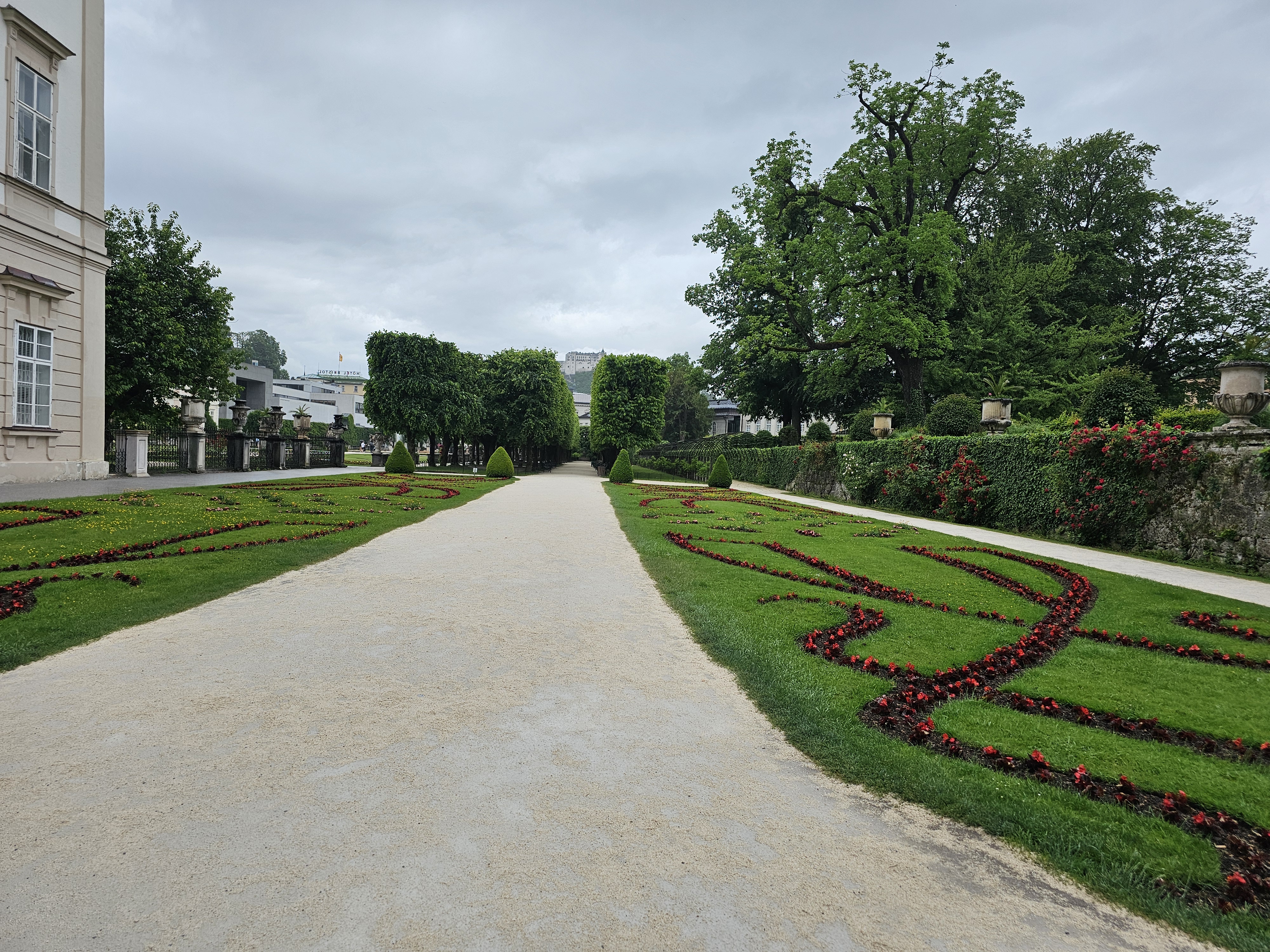 A gravel pathway leading through the Mirabell Gardens in Salzburg, Austria. On either side of the path, there are meticulously manicured lawns with decorative floral patterns and hedges. The background features trees, a stone wall with climbing plants, and a glimpse of Hohensalzburg Fortress in the distance.
