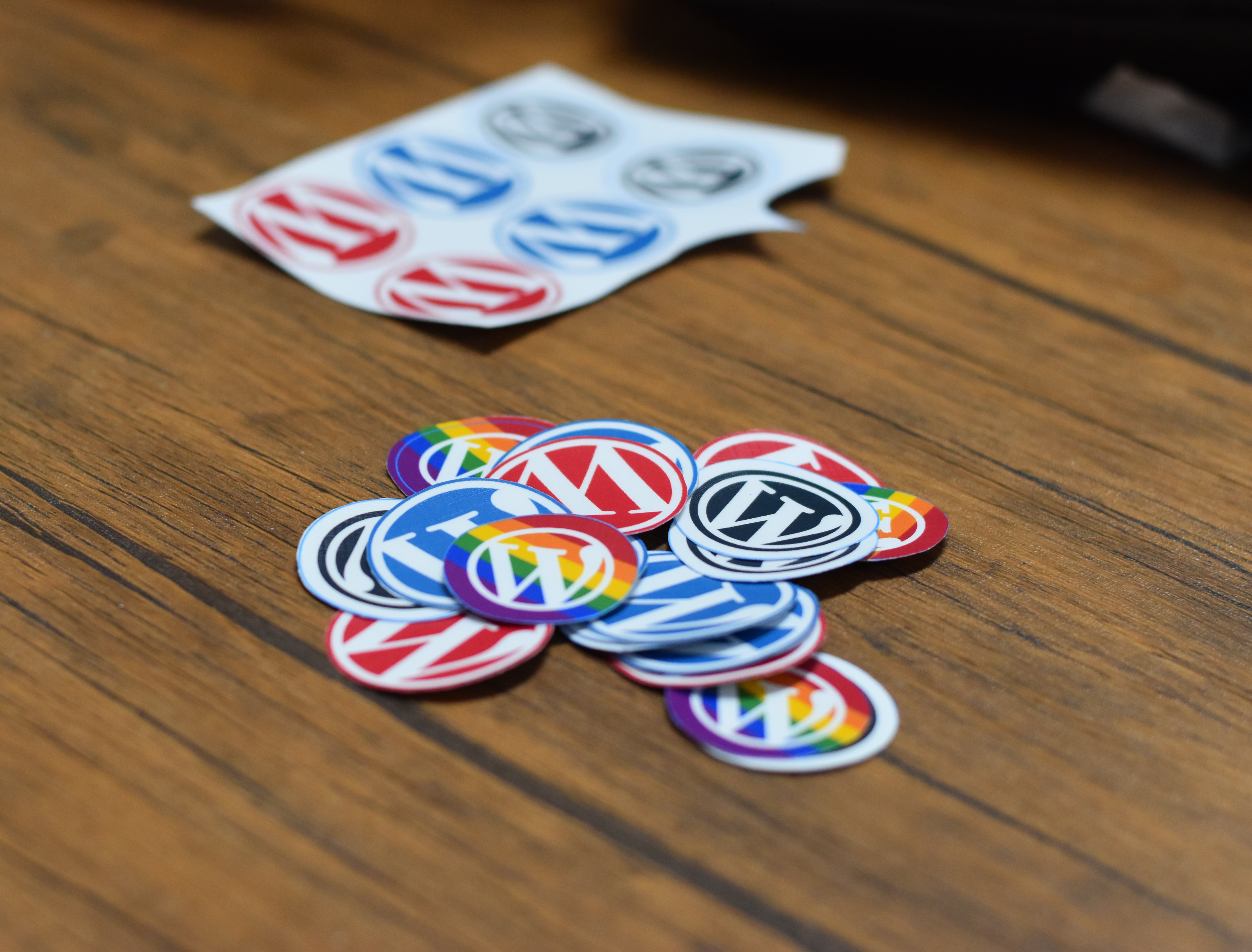 "Captured during the Iligan WordPress Meetup, a pile of colorful WordPress logo stickers—featuring red, blue, black, and rainbow-themed designs—lies scattered on a wooden surface. In the background, a partially used sticker sheet with similar designs is visible.