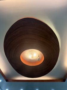 A modern ceiling light fixture integrated into a circular, wooden panel design. The fixture features a central bulb with a decorative cover and is surrounded by recessed lighting in a blue-tinted ceiling.
