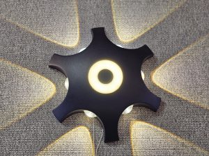 A black, star-shaped wall lamp mounted on a textured gray surface. The lamp features a central circular light and four additional lights emanating from its points, creating a pattern of light and shadow on the wall. Captured from an electrical shop in Kozhikode.
