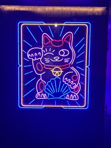 A vibrant neon sign depicting a Japanese lucky cat, known as Maneki-neko, with one paw raised. 