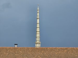 A long view of the spire of the Mole Antonelliana in Turin (Torino), Italy, rising above a terracotta-tiled roof against a cloudy sky. The architectural details of the spire are visible, showcasing its tiered design and ornamentation. 
