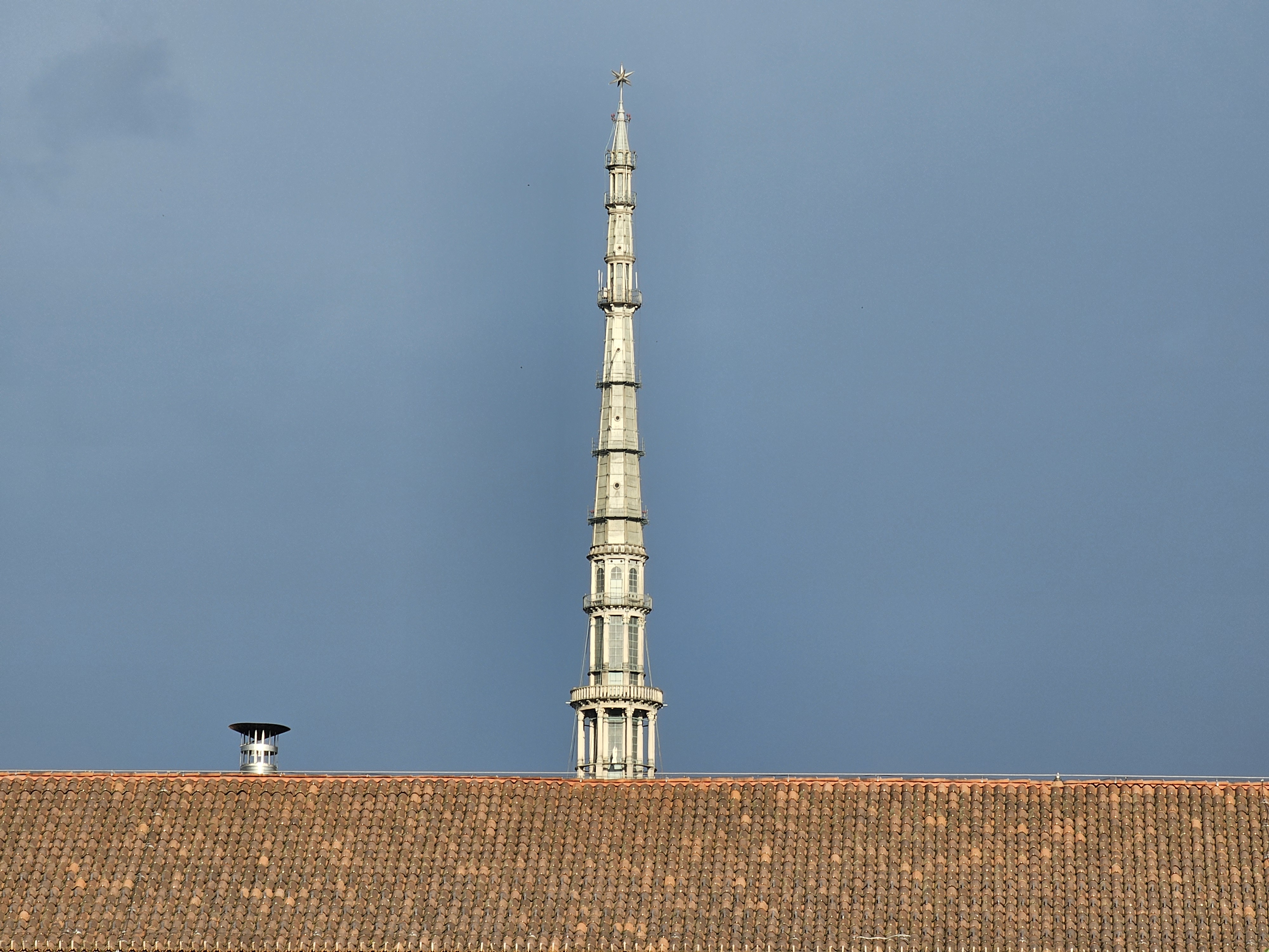 A long view of the spire of the Mole Antonelliana in Turin (Torino), Italy, rising above a terracotta-tiled roof against a cloudy sky. The architectural details of the spire are visible, showcasing its tiered design and ornamentation.
