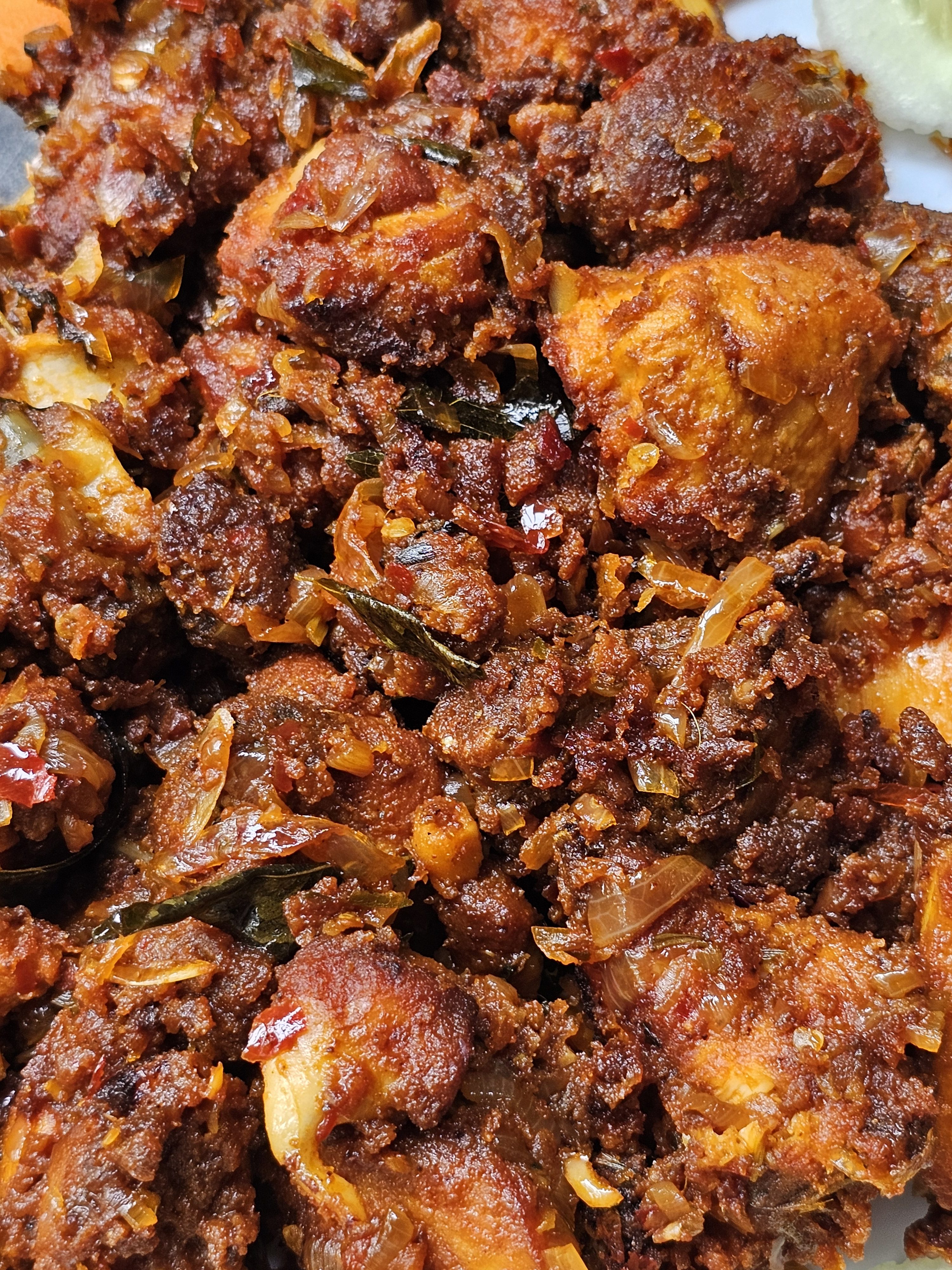 A close-up of Mangalorean Chicken Sukka, also known as Chicken Kondattam, featuring pieces of chicken coated in a rich, dark, and spicy masala, interspersed with sautéed onions and curry leaves. The dish is fried to a crisp texture, with a deep reddish-brown color indicating the use of vibrant spices. Spotted in Kozhikode, Kerala.