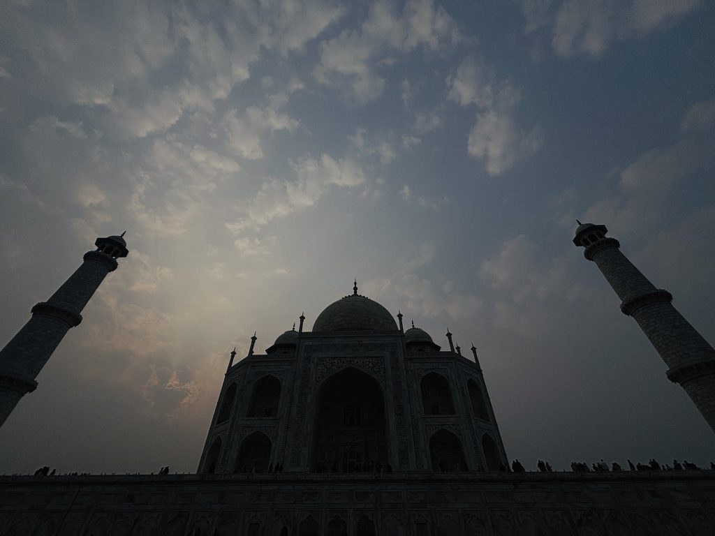 Silhouette of the Taj Mahal at sunset with cloudy skies and two minarets on either side.