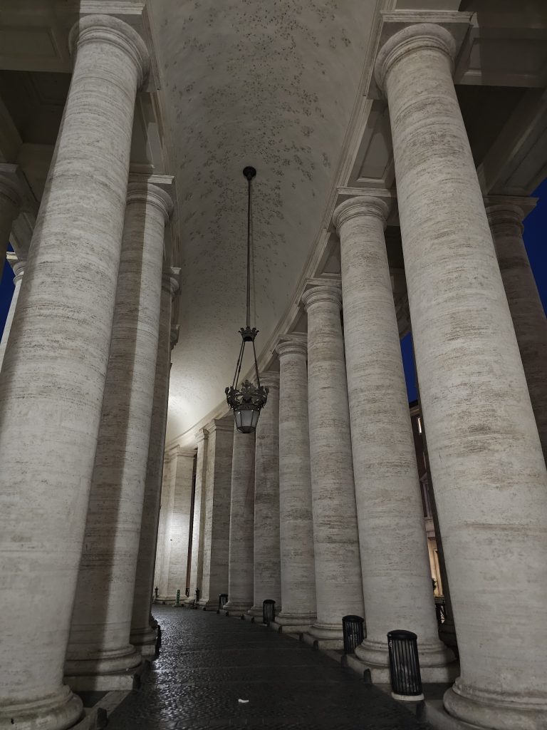 A colonnade with tall, white columns stretching into the distance, it is part of St. Peter’s Square in Vatican City. A hanging lantern provides light.