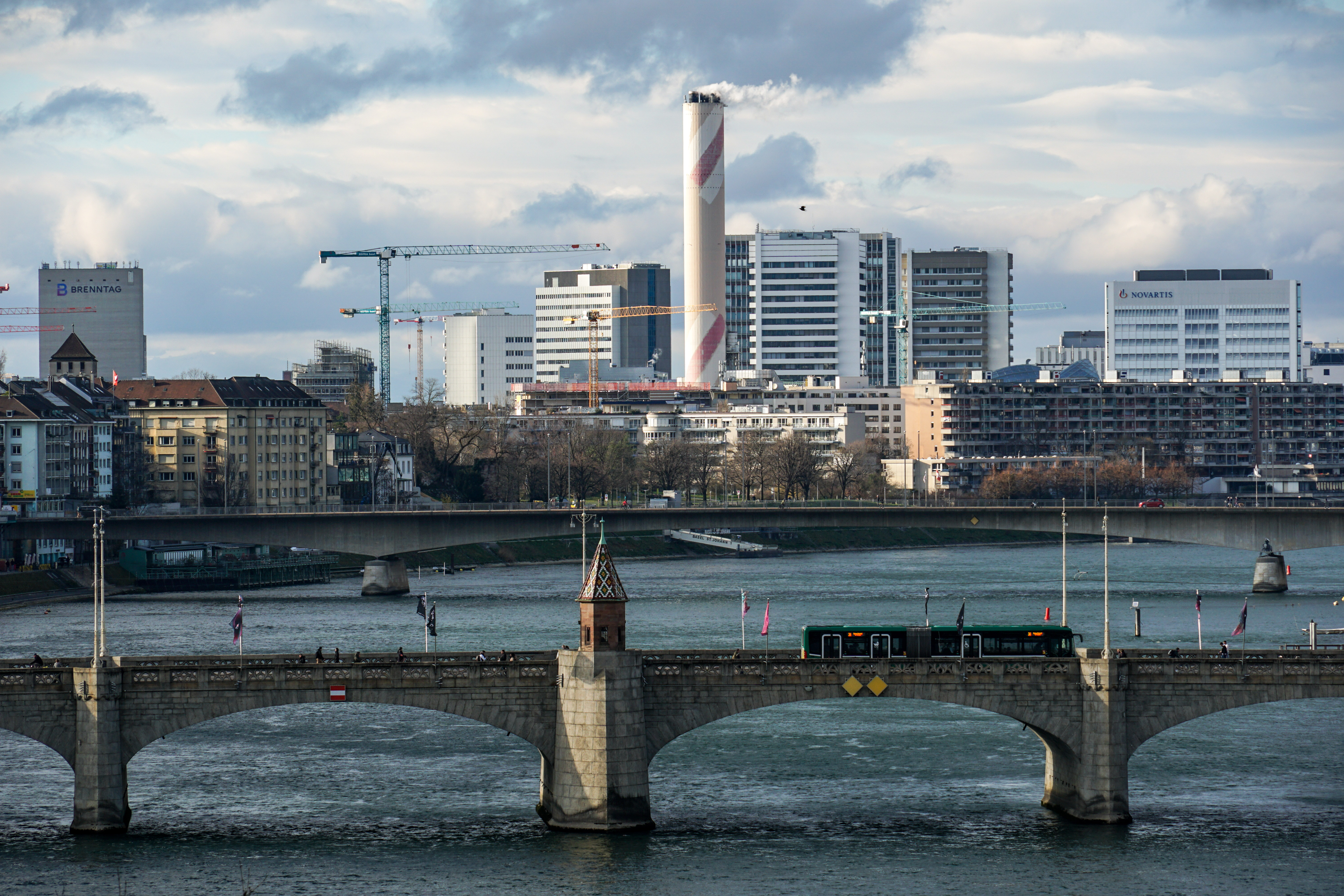 A cityscape featuring a bridge over a river with a tram crossing it. In the background, there are modern buildings and cranes indicating construction activity. The scene is under a cloudy sky.