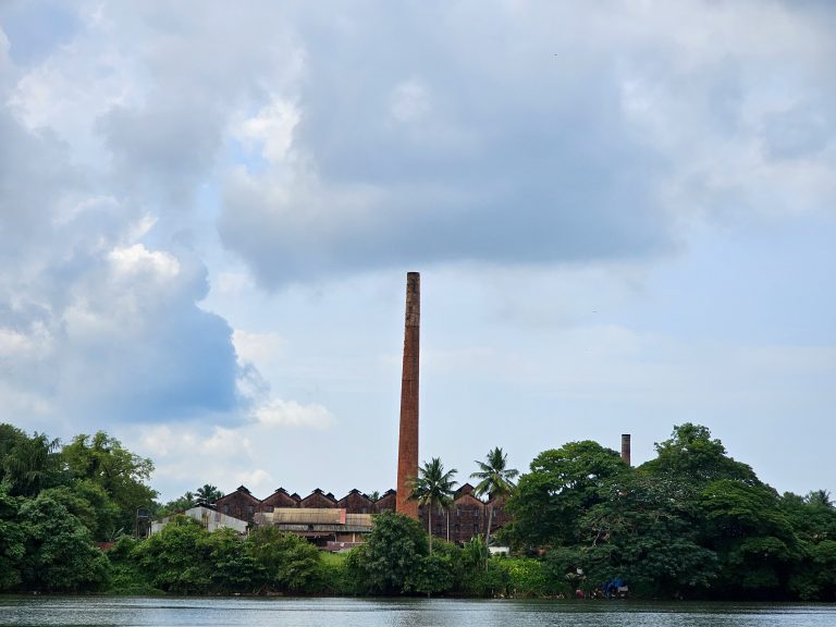 A tile factory on the banks of Chaliyar river in Feroke, Kerala, with its distinctive tall chimney. The factory buildings, characterized by their red-tiled roofs, are partially obscured by lush green trees and vegetation. The chimney, made of red brick, stands prominently against a backdrop of a cloudy sky.