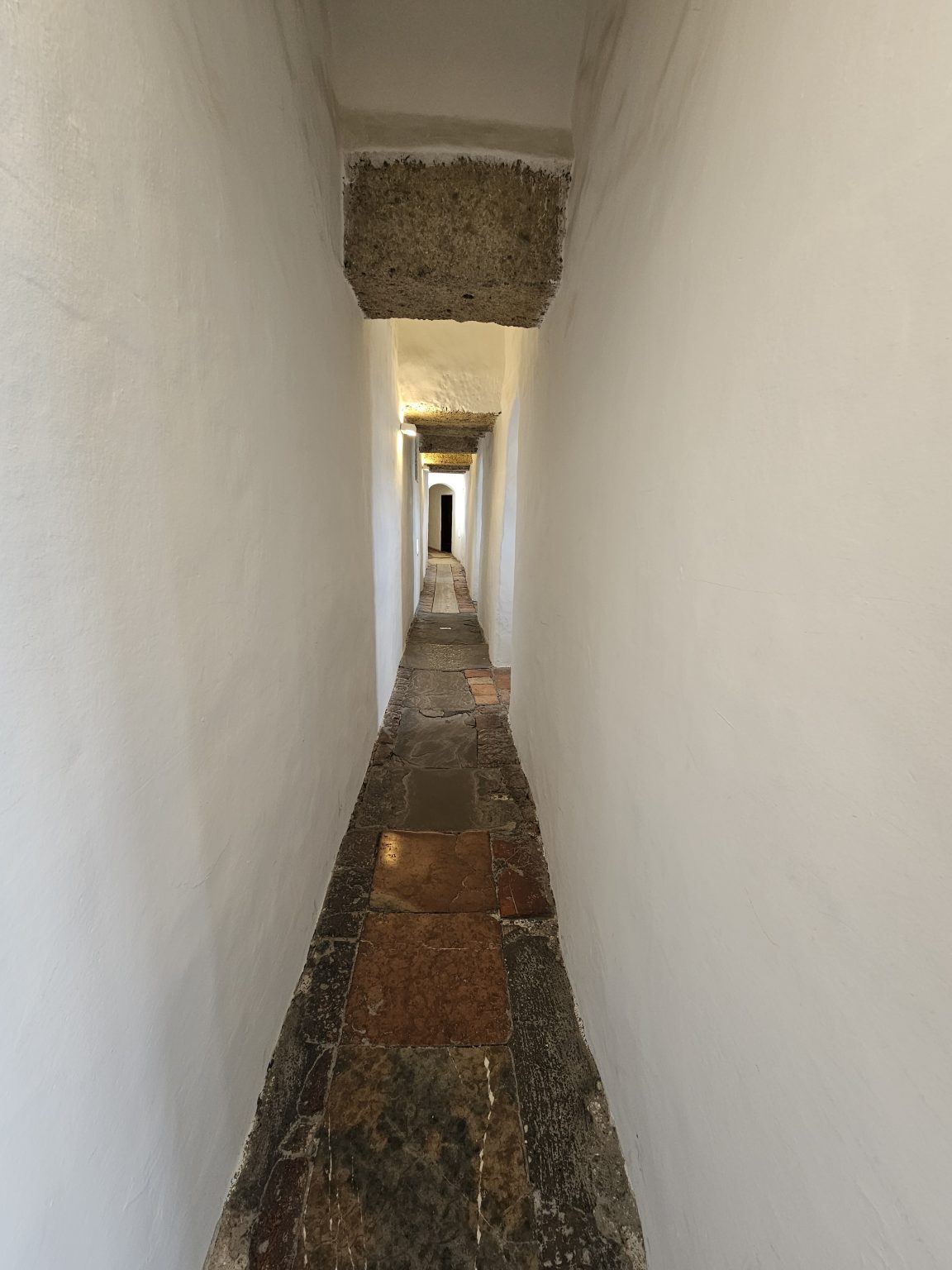 A long, narrow passageway with white walls and a stone floor. The lighting is dim, with the main source of light coming from the far end of the corridor, highlighting the depth and length of the space. Captured from Hohensalzburg Fortress, Salzburg, Austria.