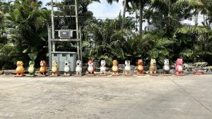 A row of colorful cartoon-like animal statues is lined up on a street with lush greenery and palm trees in the background. There is a variety of animal figures, each with distinct colors and patterns.
