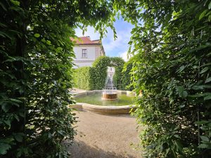 A fountain of Waldstein Garden in Prague, framed by lush green foliage. The fountain is situated in a circular basin with a stone edge, and the water feature has a tiered design. The area around the fountain is covered in light-colored gravel. The view is framed by an archway of green bushes, creating a natural border at the edges of the image. 