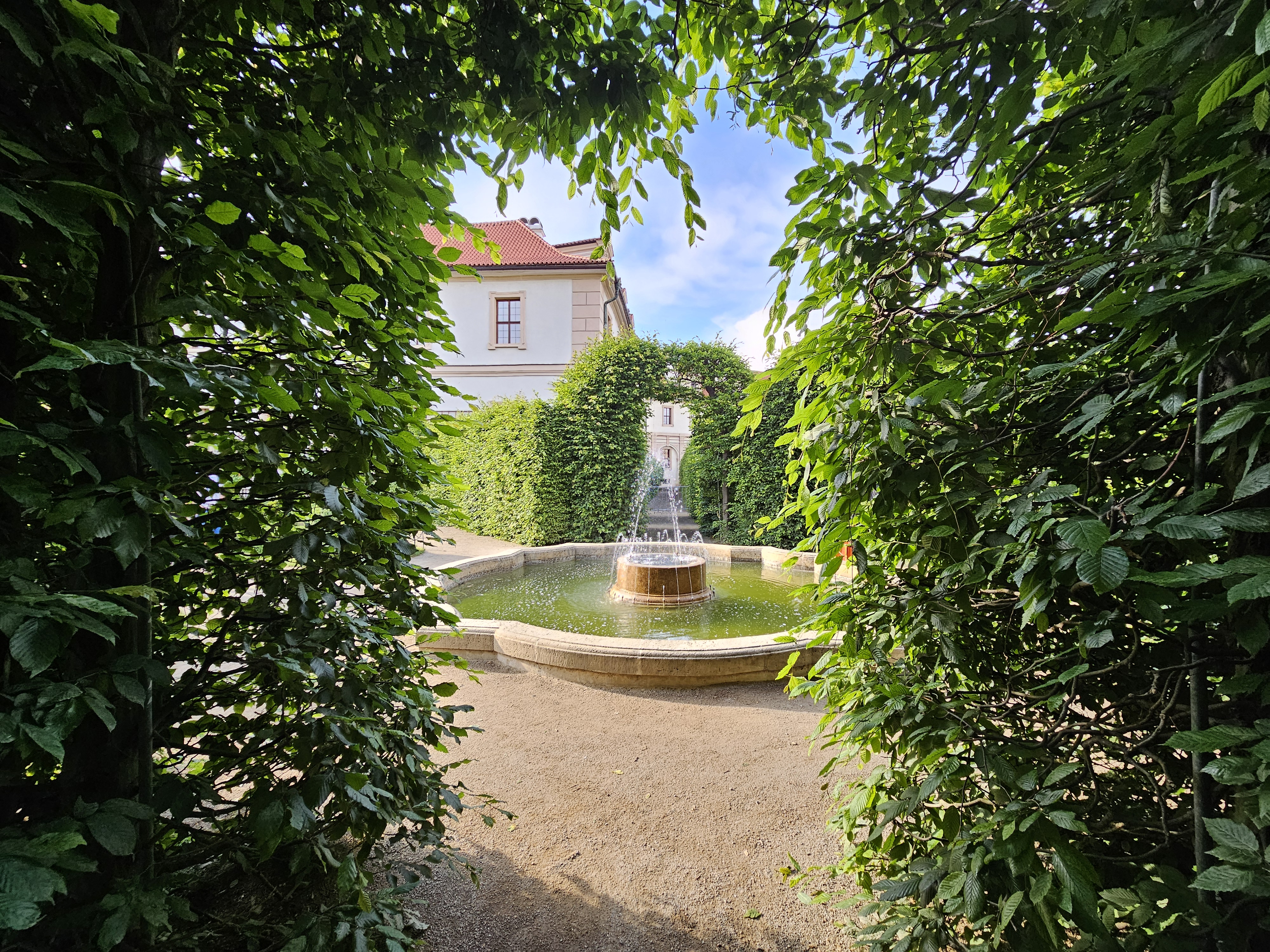 A fountain of Waldstein Garden in Prague, framed by lush green foliage. The fountain is situated in a circular basin with a stone edge, and the water feature has a tiered design. The area around the fountain is covered in light-colored gravel. The view is framed by an archway of green bushes, creating a natural border at the edges of the image.
