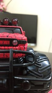 A close-up view of a red toy monster truck with a cracked paint design, featuring large black tires and a front grille.
