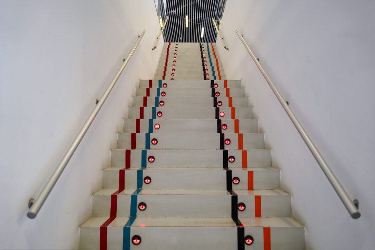 A staircase with white steps, marked by multicolored vertical lines in red, blue, orange, and black. Each step features a small red light between the lines. The staircase is bordered by stainless steel handrails and leads to an area with a ceiling of vertical black slats.