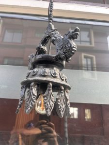 A decorative, ornate black metal light fixture with dragon design elements and leaf-like details hangs in front of a window. A clear light bulb is visible beneath the fixture. Reflections of buildings and a person taking a photo are seen on the glass.