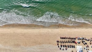 Aerial view of Cavelossim Beach in South Goa, showing a shoreline where ocean waves meet the golden sand. Rows of sun loungers and umbrellas are neatly arranged near a beachside shack. A few people are visible walking along the shore and in the water, while tire tracks crisscross the sandy beach.