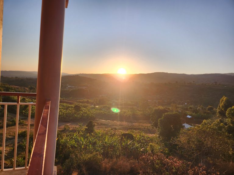 A scenic view of a sunrise over rolling hills and lush greenery, as seen from a balcony with a railing in the foreground.