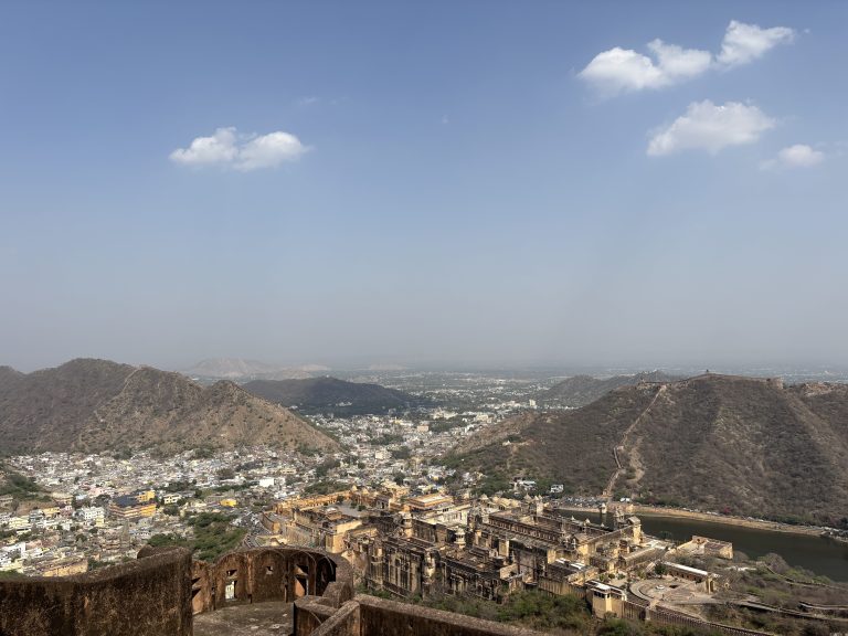 Aerial view of a historic fort complex surrounded by a dry, hilly landscape. The fort features large, ornate buildings with courtyards and towers. Below the fort, a city is visible, nestled among the hills. A few fluffy clouds are scattered in the clear blue sky.