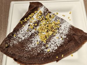 A folded, triangular chocolate crêpe, dusted with powdered sugar and sprinkled with chopped pistachios. The crêpe is presented on a white, square plate. Captured from a restaurant in Turin, Italy.  
