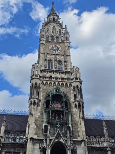 A Gothic-style clock tower with intricate carvings and a large clock face, set against a partly cloudy blue sky. There are decorative elements and statues on the facade.